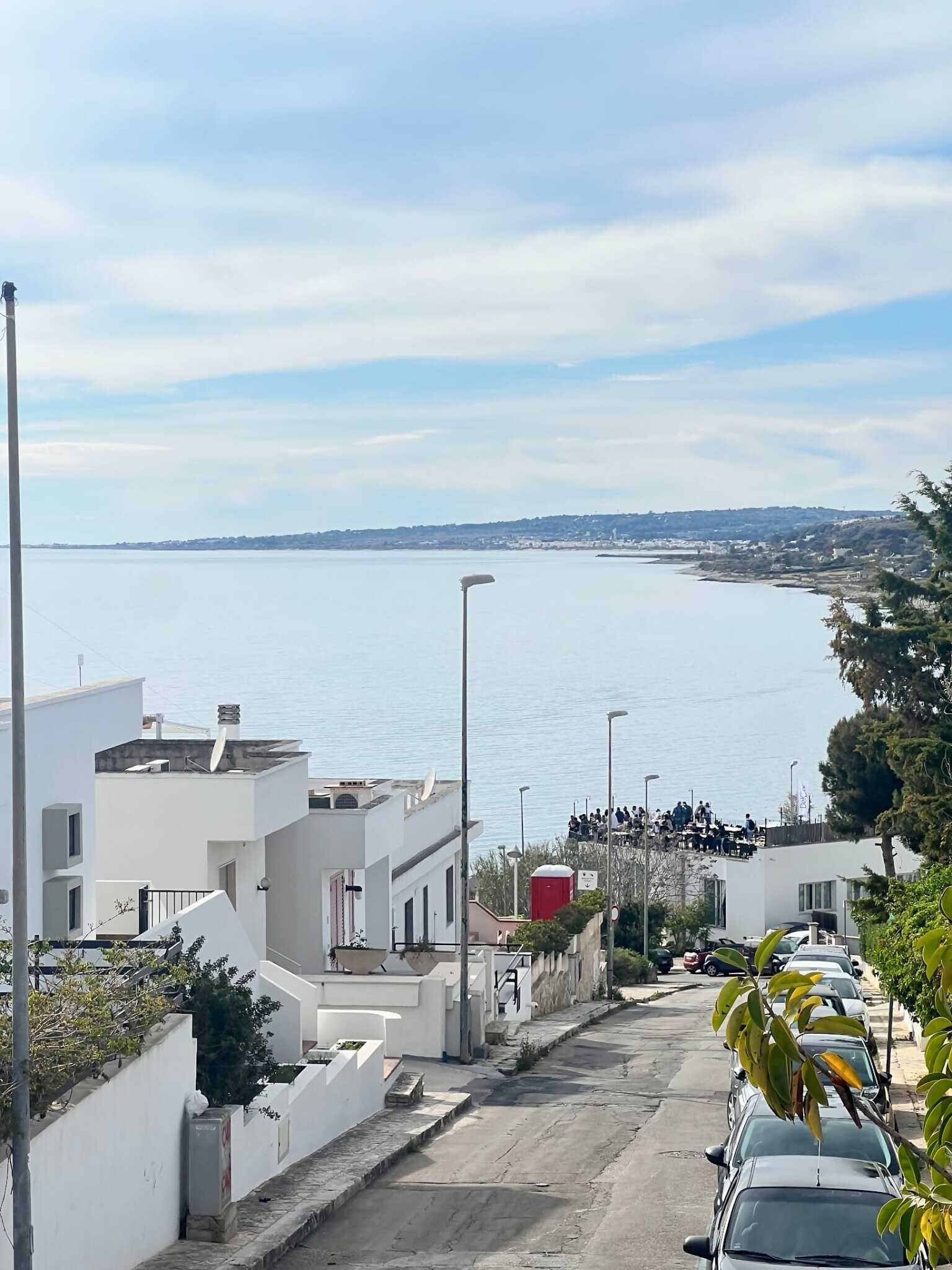 Una calle costera con edificios modernos, una vista despejada del mar y coches aparcados.