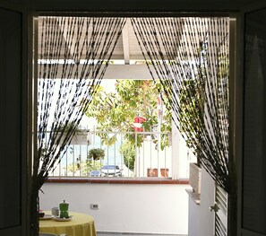 A room with a view through a window with a white curtain, a table with a yellow tablecloth, and a potted plant on the windowsill. - Villa with sea view in San Gregorio - Patu (Patu')