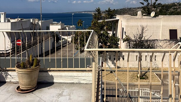 A rooftop with a potted plant, a metal railing, and a view of the sea and distant buildings. - Villa with sea view in San Gregorio - Patu (Patu')