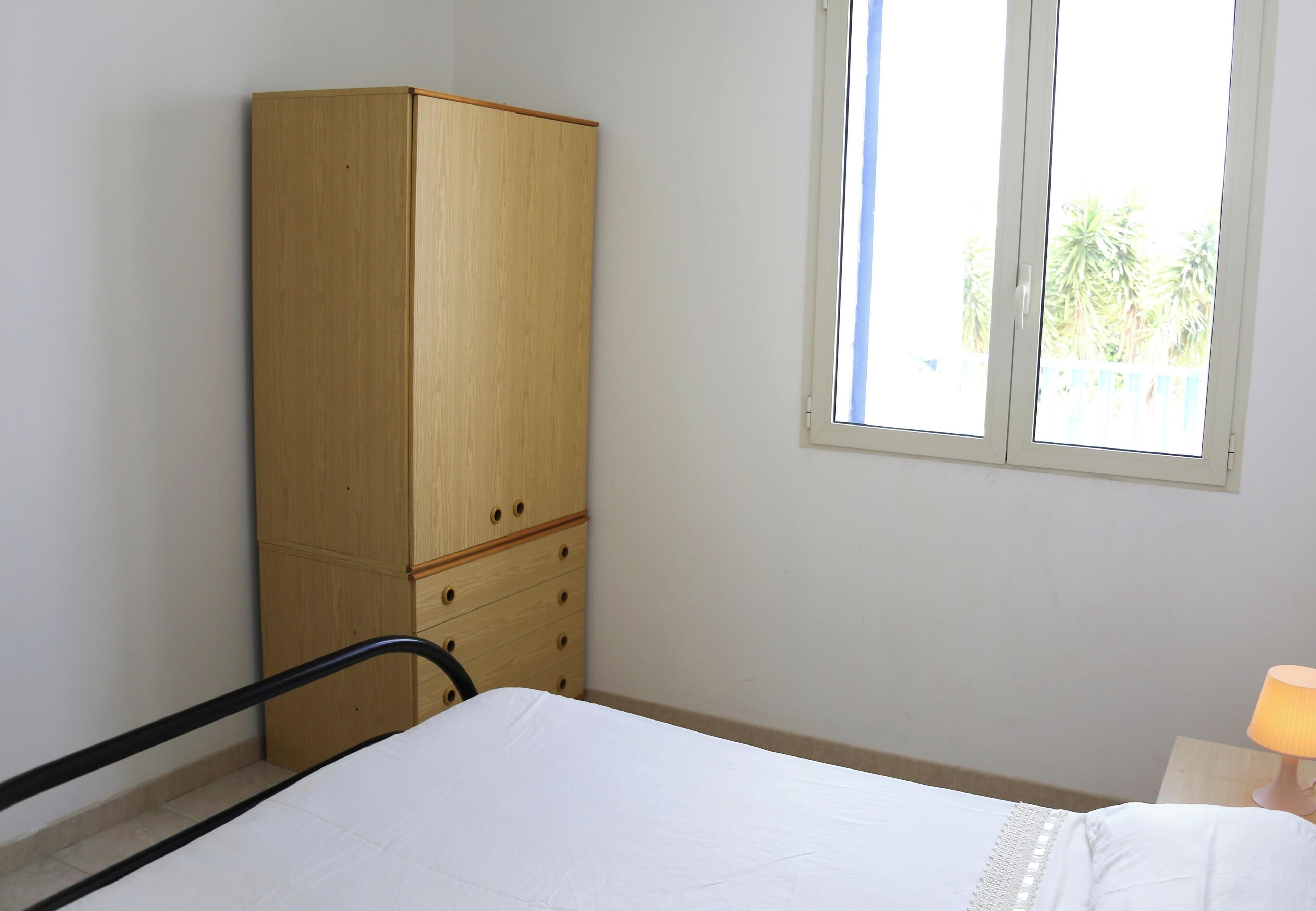A bedroom with a bed, a wooden wardrobe, a window, and a nightstand with a lamp.