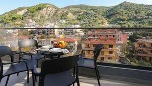 Un balcón con mesa lista para comer, sillas y un bol de fruta. La vista incluye una colina con edificios y un cielo despejado.