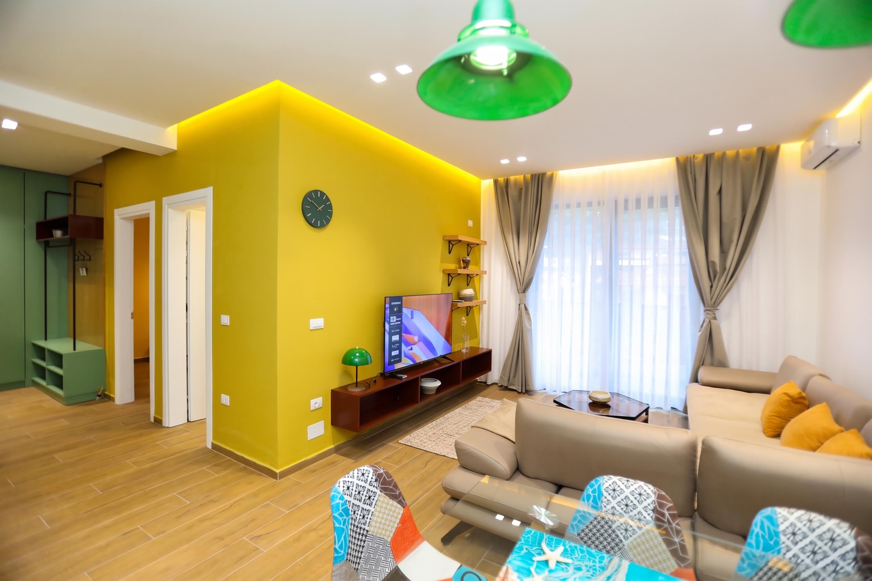 A modern living room with a yellow accent wall, a flat-screen TV, a sofa, and a dining area with patterned tableware.