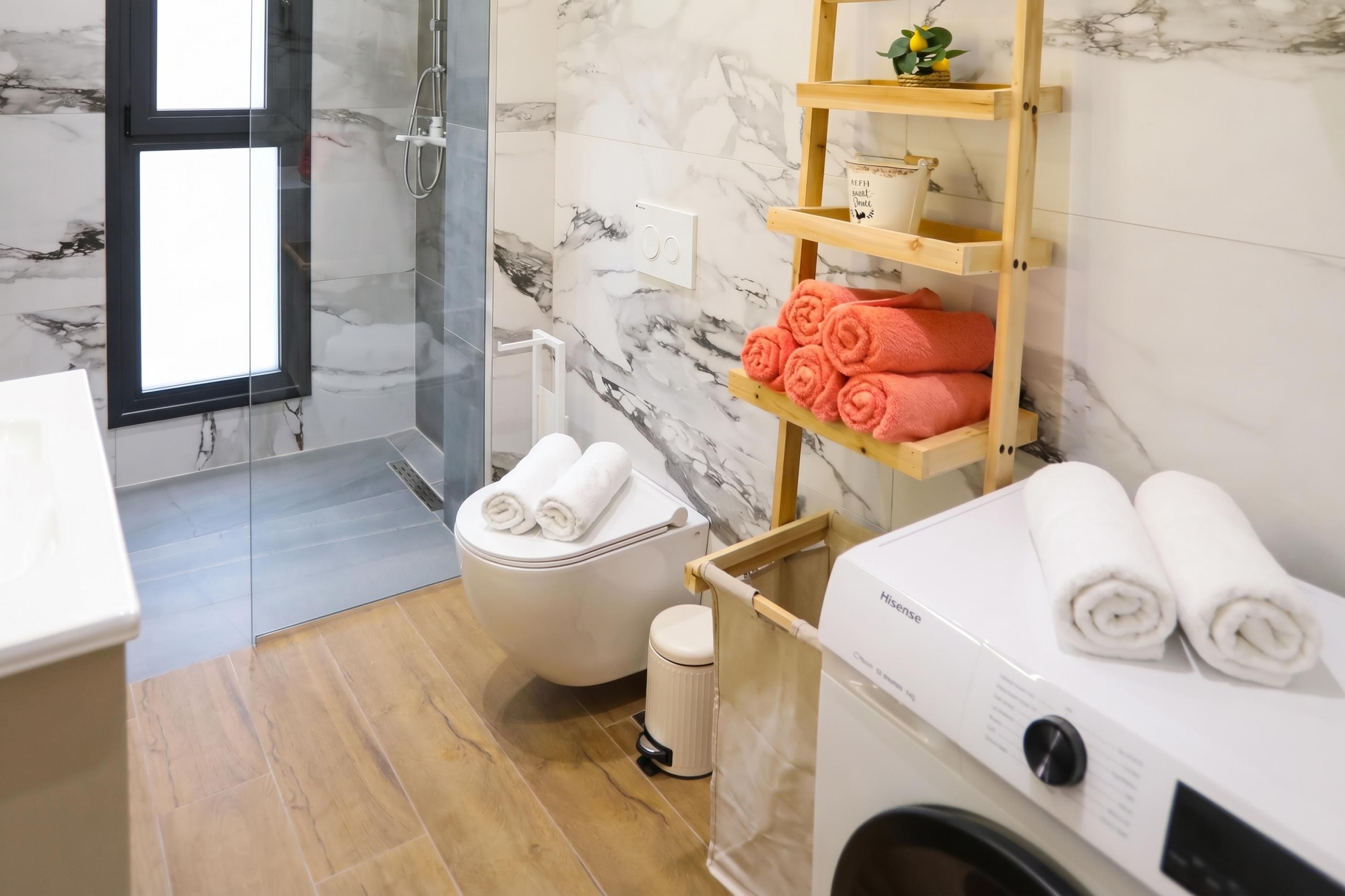 A modern bathroom with a glass-enclosed shower, a toilet, a washing machine, and a shelf with towels.