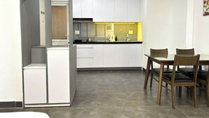 A compact kitchen with white cabinets, a dining table with chairs, and a small refrigerator. - Nghe Home (Da Nang)