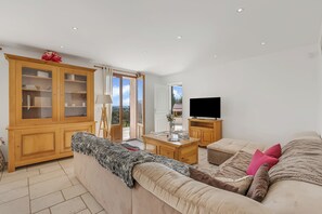 Een ruime woonkamer met een bank, televisie, houten kast en uitzicht door de deur.