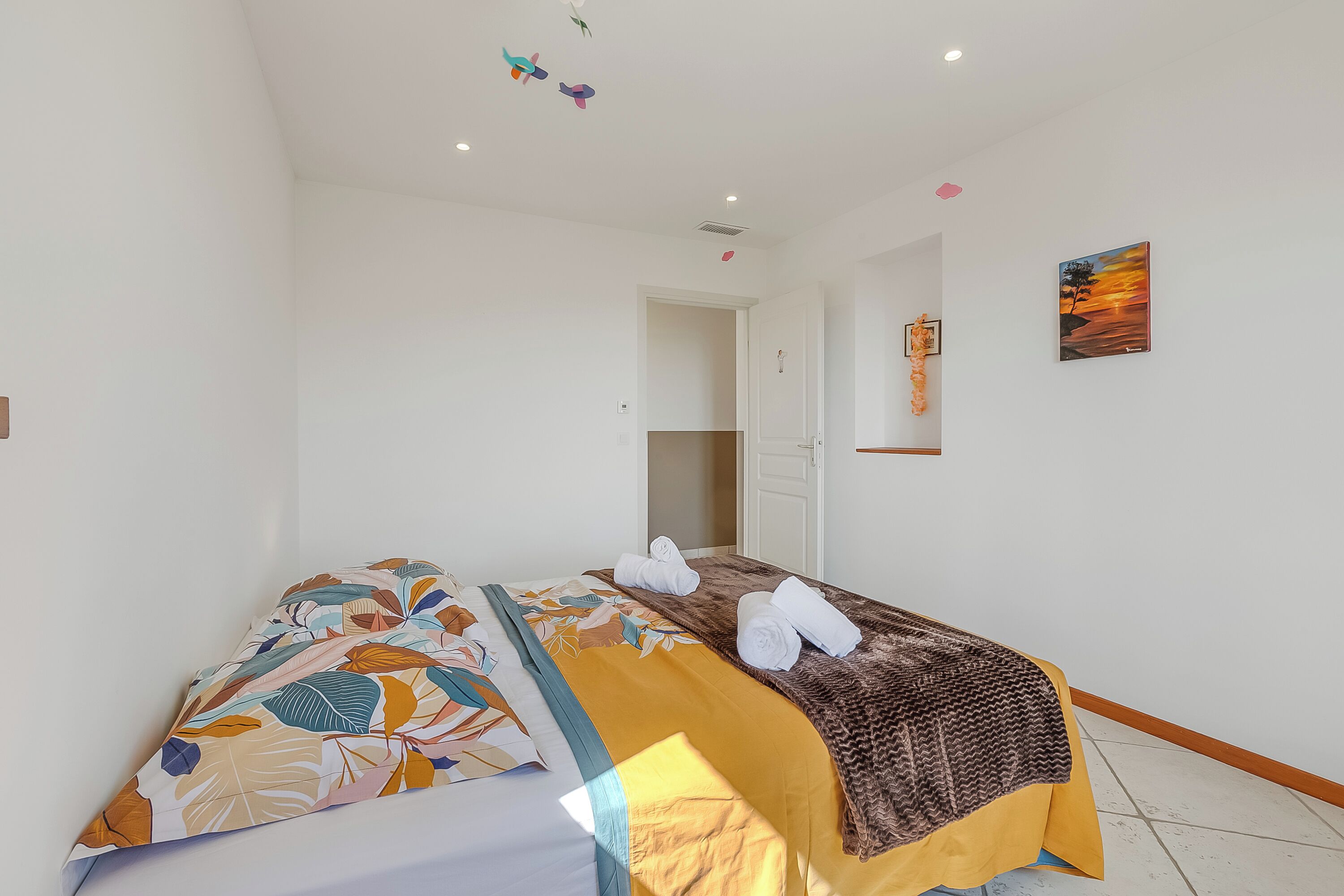 A bedroom with a bed, two towels, a painting, and a door leading to another room.