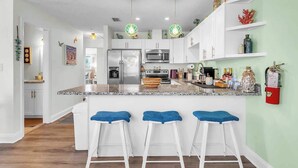 A modern kitchen with white cabinets, a granite countertop, and three bar stools with blue cushions. - Beach in 90 Seconds w/ Hot Tub, Coffee Bar & Games (Indian Rocks Beach)