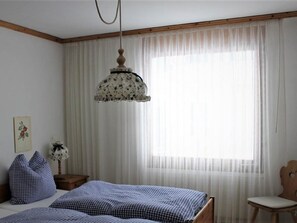 A bedroom with a bed, a chair, a nightstand, a hanging lamp, and a window with sheer curtains. - Chesa Piz Alv - Roseg Spirit by Interhome (Pontresina)