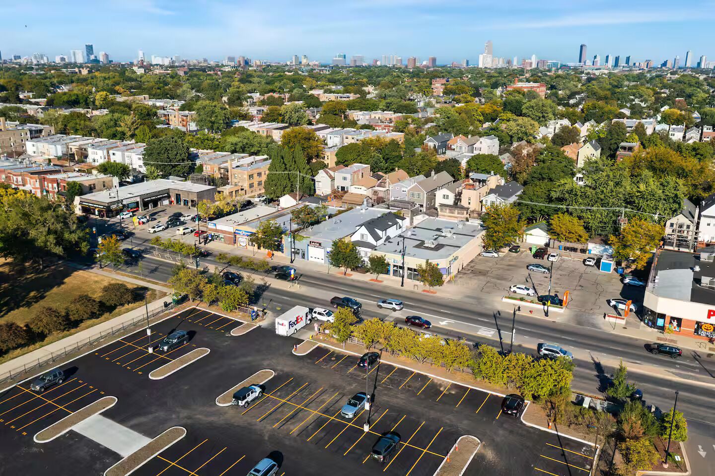 An aerial view of a cityscape with a mix of residential and commercial buildings, a parking lot with several cars, and a clear sky.