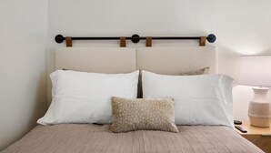 A bed with two white pillows and a patterned pillow, a headboard with a wooden horizontal bar, and a bedside table with a lamp and a remote control.