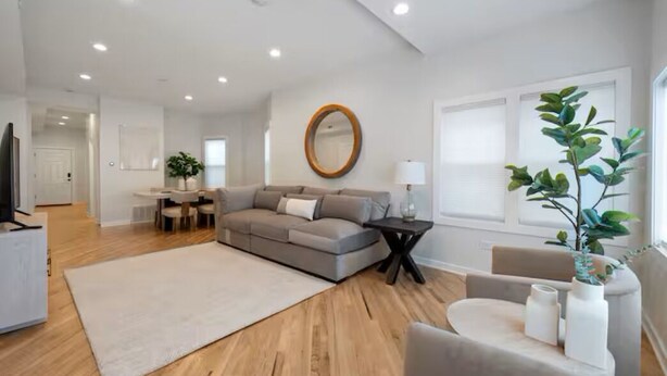 A modern living room with a sofa, a coffee table, a TV, a dining area, and a large window with a plant.