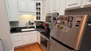 Eine Küche mit weißen Schränken, einem Edelstahlkühlschrank, einem roten Standmixer und einem gemusterten Teppich.