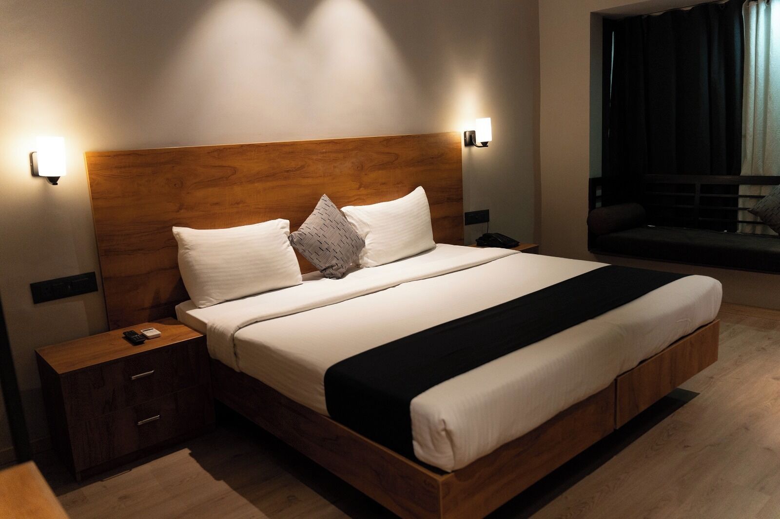 Een moderne hotelkamer met een houten hoofdeinde, een groot bed met wit beddengoed, een nachtkastje met een afstandsbediening.