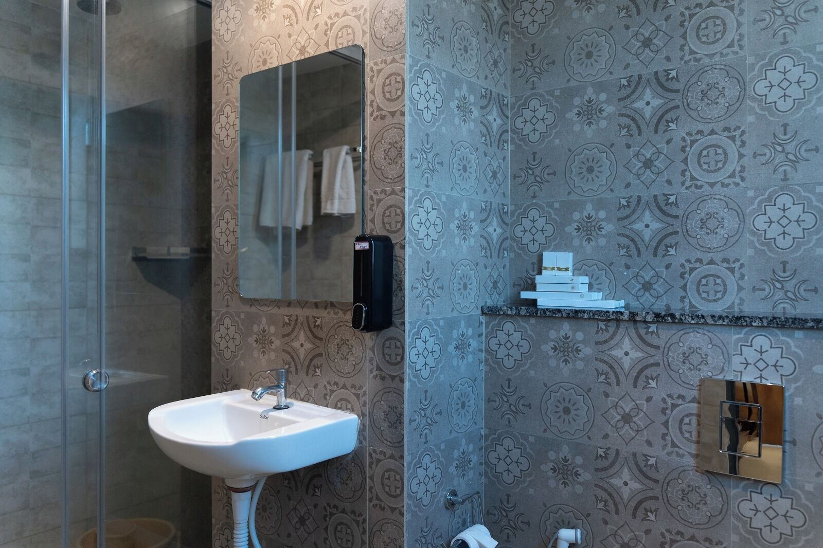 A bathroom with a glass shower enclosure, a white sink, a mirror, patterned wallpaper, and a shelf with toiletries.