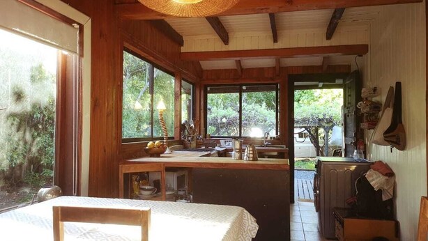A rustic cabin with a wooden interior, a dining area with a table and chairs, a kitchen with a sink and counter, and large windows offering views of the surrounding forest. - Casa del Estero in downtown Pucón (Pucon)