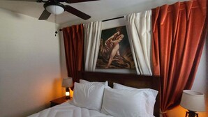 Une chambre d’hôtel avec un lit, un ventilateur de plafond, un tableau et deux lampes de chevet.