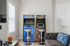 A room with two arcade machines, a couch with a blue pillow, and a window with blinds.