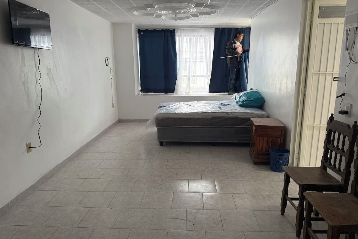 A room with a bed, a wooden chair, a small table, a window with curtains, and a ceiling fan.