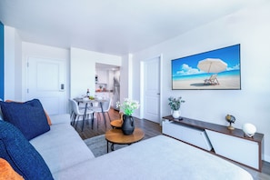 A modern living room with a sofa, a coffee table, a TV, and a dining area in the background. - Luxury Downtown Fort Myers Apt/Pool View • King Bed • Walk to Dining/Nightlife (Fort Myers)