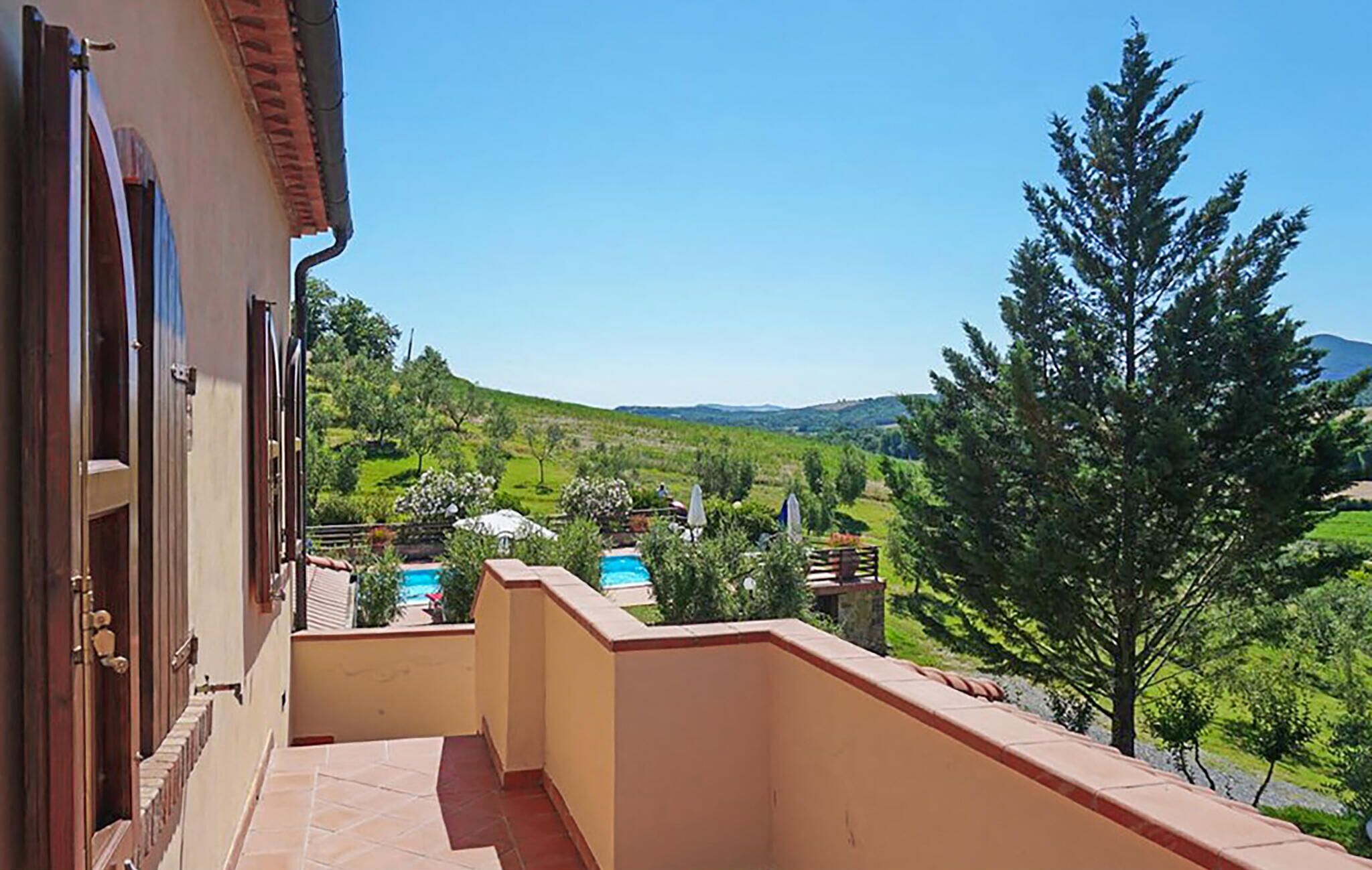 A balcony with a view of a pool and greenery, a building with a tiled roof, and a clear blue sky.