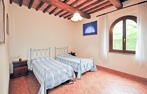 Ein Zimmer mit zwei Betten, einem Vorhangfenster, einer Holzbalkendecke und einem kleinen Tisch mit Lampe.