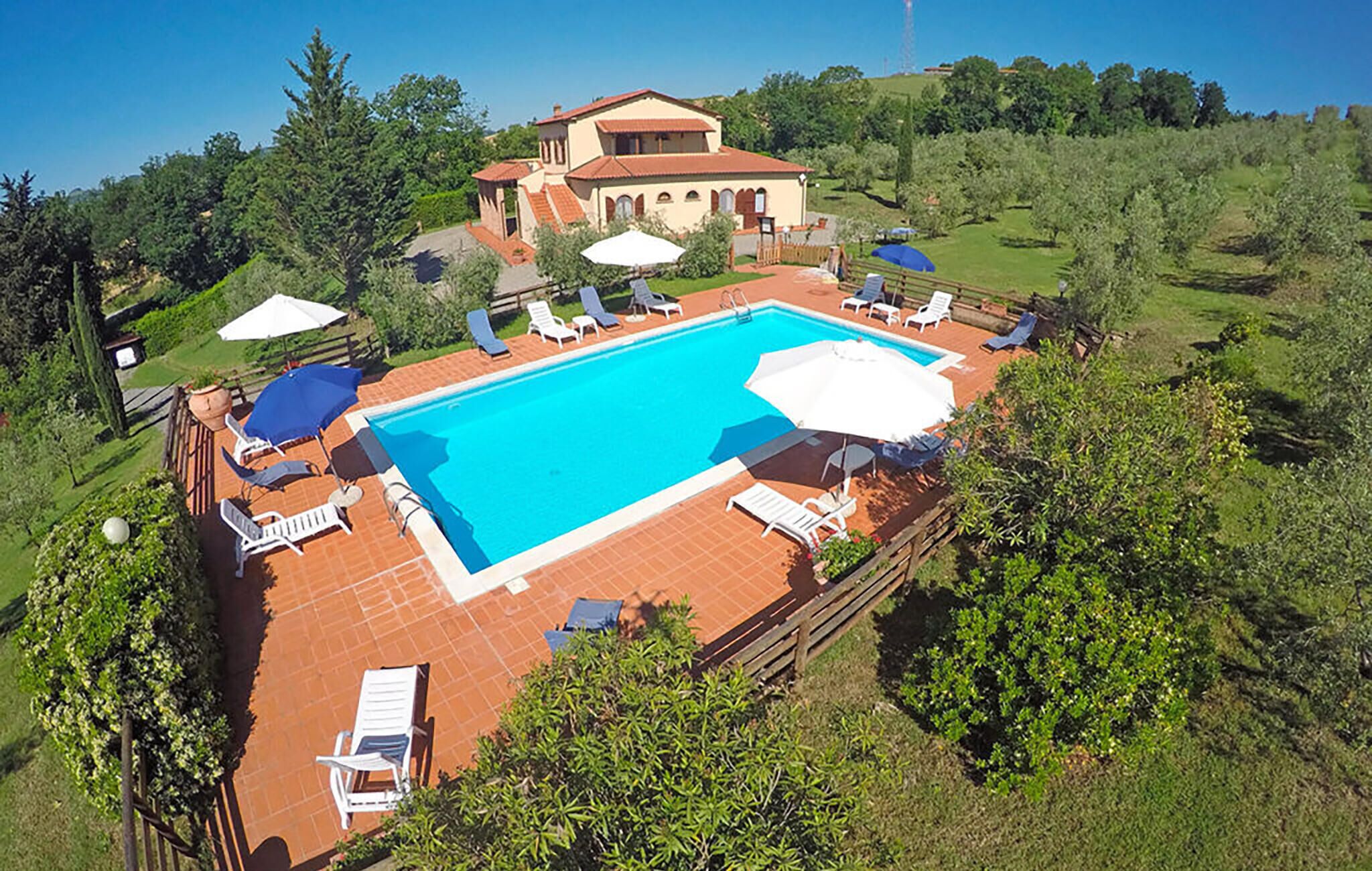 A rectangular swimming pool with lounge chairs and umbrellas, surrounded by a wooden deck and a building with a tiled roof.