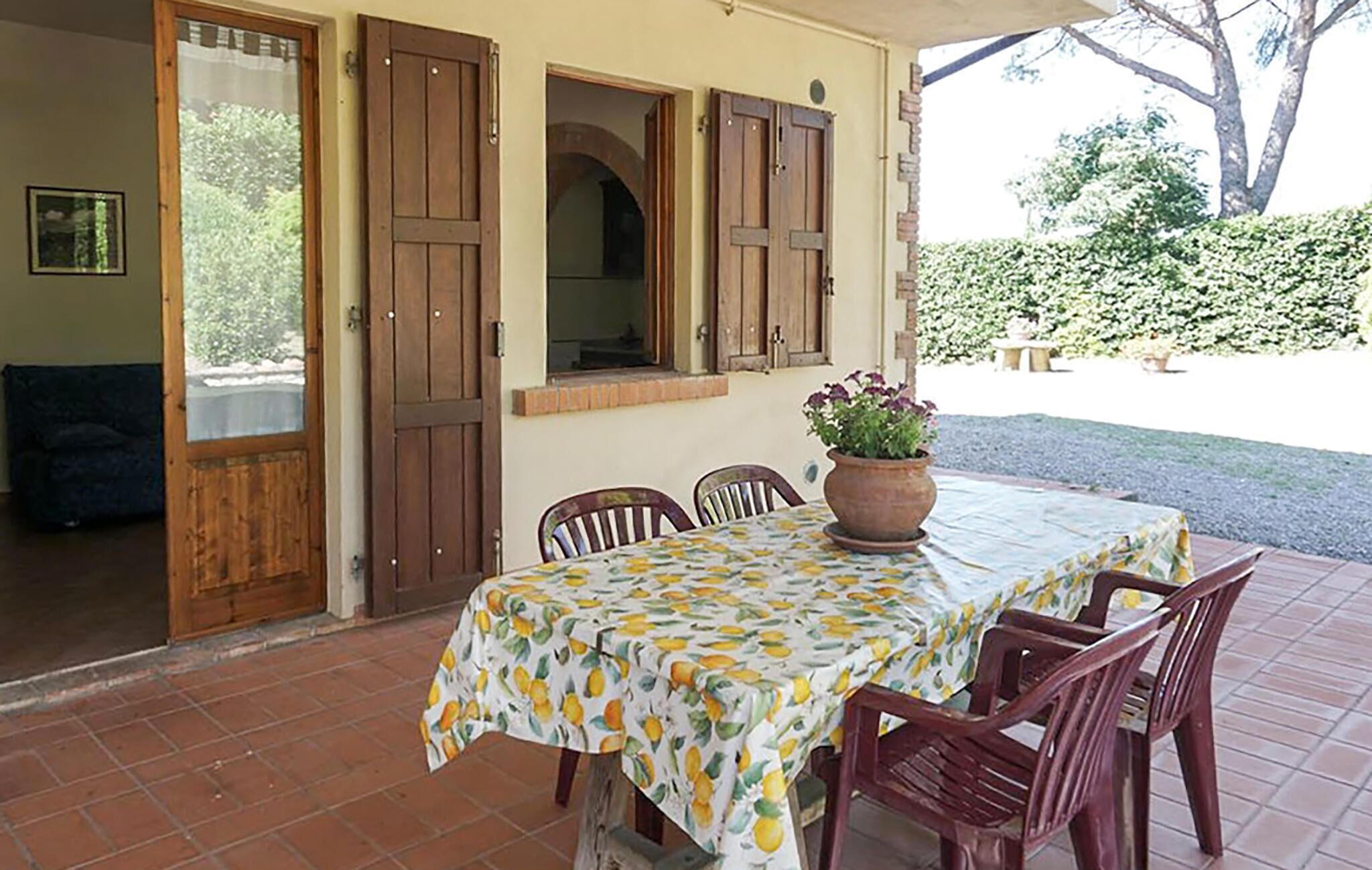 Un área de comedor al aire libre con una mesa cubierta por un mantel estampado con motivos de frutas, rodeada de sillas de madera y una planta en maceta sobre la mesa.