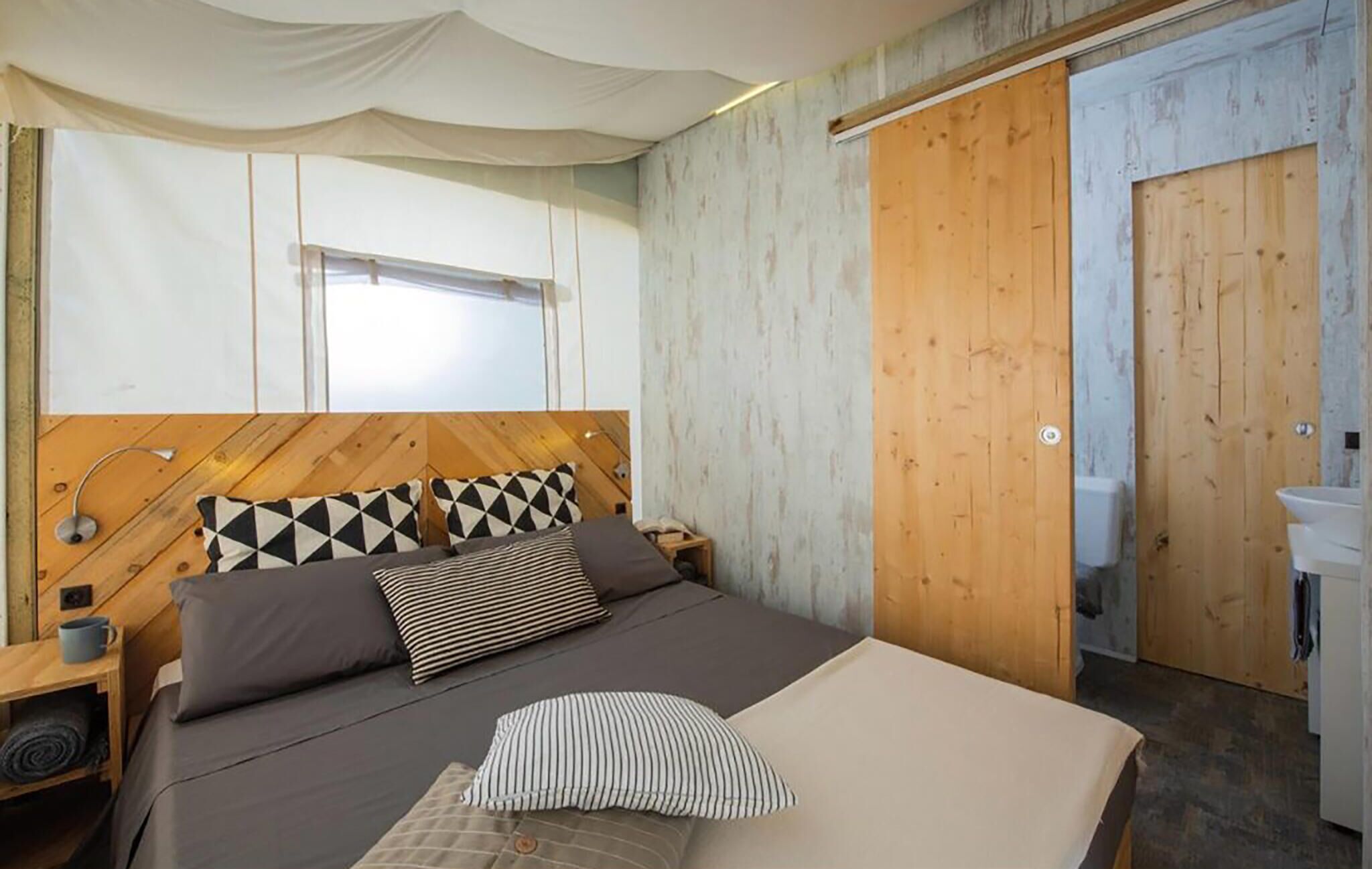 A bedroom with a bed, a bedside table, a lamp, a window, and a bathroom door.