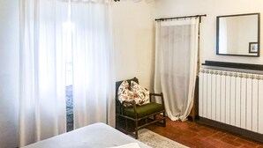 Une chambre à coucher avec un lit, une chaise recouverte d’un plaid à motifs floraux, un radiateur et des rideaux en voile.