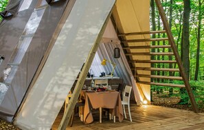 Una tenda con un'area pranzo all'interno, una scala di legno e uno sfondo boschivo.