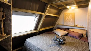 Ein Schlafzimmer mit schrägen Wänden, einem Bett mit einer gesteppten Decke, Kissen, einem kleinen Flugzeugmodell und einer Wand mit einer großen Buchstabenleiste.