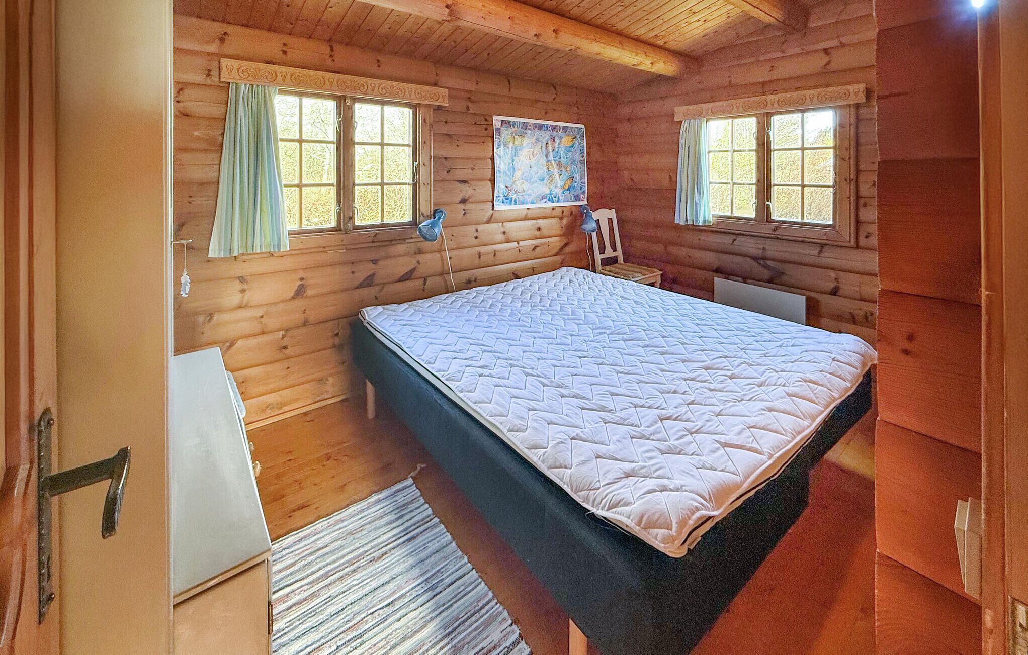 Ein Schlafzimmer einer Holzhütte mit einem Bett, zwei Fenstern, einem kleinen Tisch und einer Wandlampe.