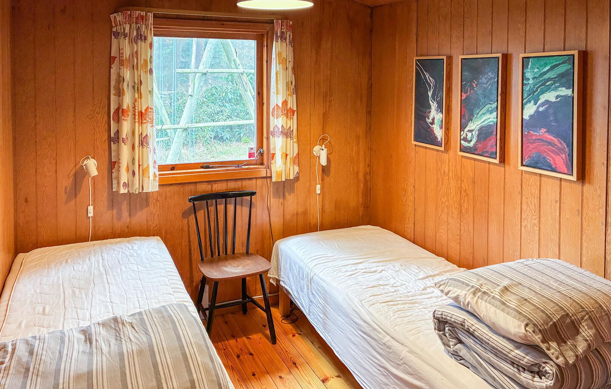 Una habitació petita amb dos llits individuals, una cadira, una finestra amb cortines i tres quadres al mur.