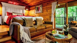 Interior acogedor de una cabaña con techo de madera, un sofá de cuero, una mesa de comedor con revistas y un ventanal con vistas a una terraza.