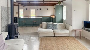 Een gezellige zithoek met een bank, een geweven vloerkleed, een klein rond tafeltje, een keuken met een barkruk en een groot raam.