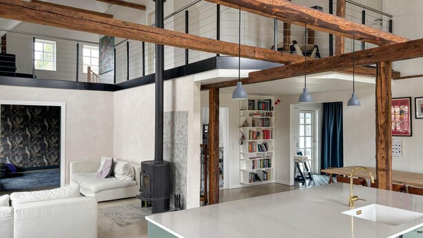 Een ruime woonkamer met een hoge plafond, zichtbare houten balken, een centraal gelegen open haard, een witte bank, een keukeneiland met een spoelbak en een boekenkast.