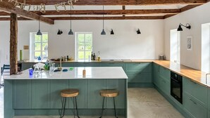 Een keuken met een groot eiland, groene kastjes, hanglampen en uitzicht op groen buiten.