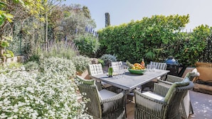 Eine Terrasse im Freien mit Korbmöbeln, einem Tisch gedeckt mit Gläsern und einer Obstschale, sowie einem Grill im Hintergrund.