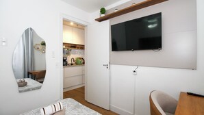 Una habitación compacta con cama, televisor en la pared, una pequeña zona de estar y una cocina al fondo.