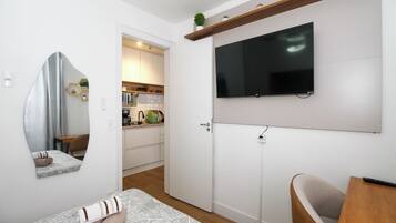 Quarto compacto com cama, TV fixada na parede, um pequeno espaço de estar e cozinha ao fundo.