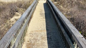 Ein hölzerner Steg führt zum Strand, begleitet von Dünen an beiden Seiten.