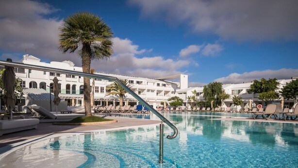 A pool area with lounge chairs, a palm tree, and a building with arched windows in the background. - 1 Bedroom Junior Suite with kitchen, Pool and Garden Views, Full Bathroom, 40sq Metres, Balcony, Lar (Corralejo)
