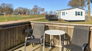Een terras met twee stoelen en een tafel, een barbecue en een stacaravan op de achtergrond.