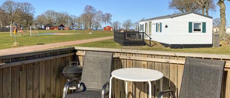 A deck with two chairs and a table, a grill, and a mobile home in the background.