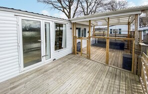 En takförsedd uteplats med trägolv, glasdörrar och utsikt över ett vitt, flyttbart hus i bakgrunden.