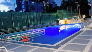En swimmingpool med ett kaklat poolområde, metallräcken och ett grönt staket i bakgrunden.