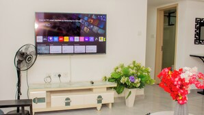 Ett modernt vardagsrum med en platt-TV som är monterad på väggen, ett soffbord med glasskiva, ett vitt konsolbord med lådor, en golvfläkt och en vas med blommor.