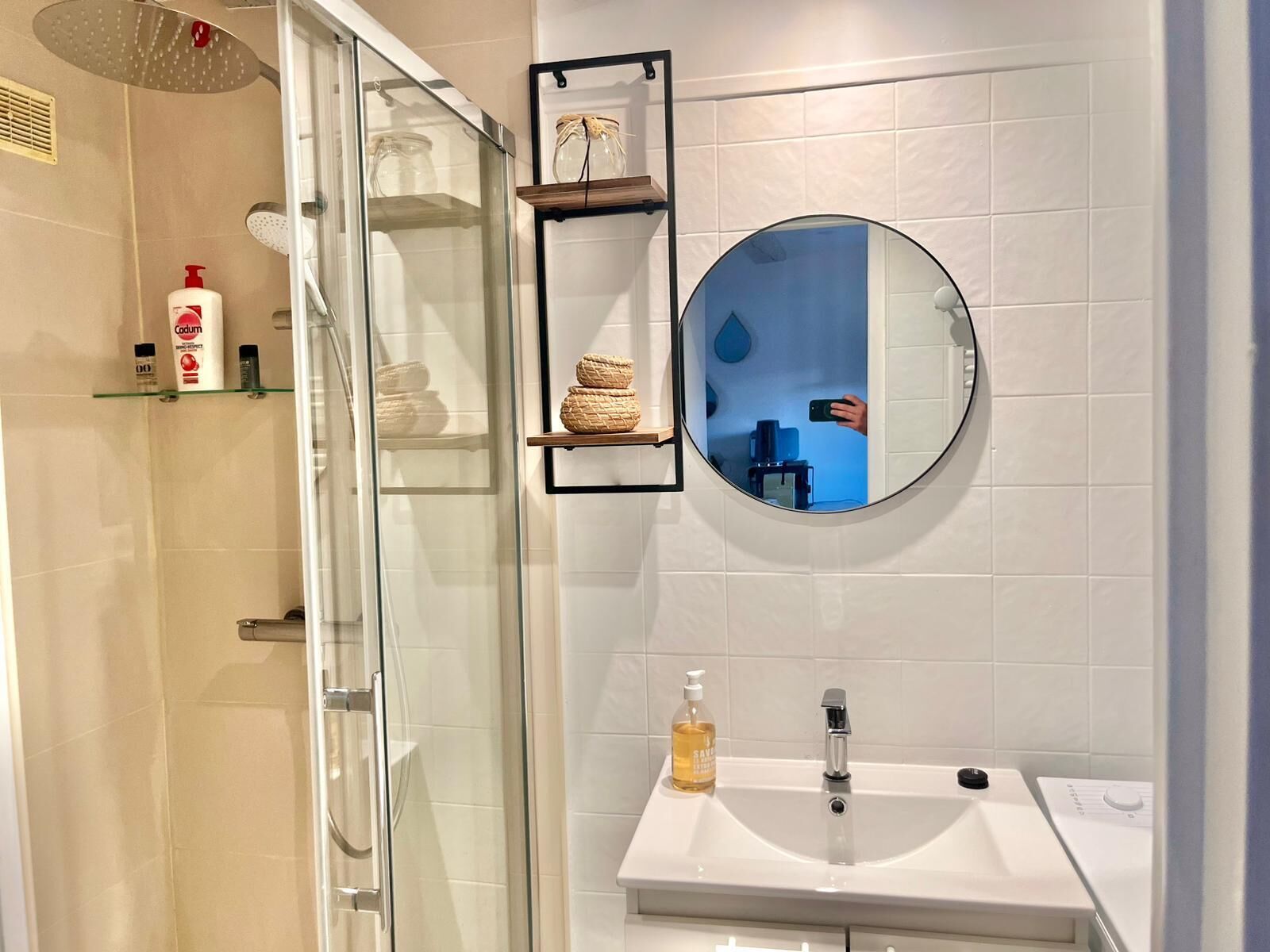 Een moderne badkamer met een glazen douchecabine, een ronde spiegel, een witte wastafel en een plank met toiletartikelen.