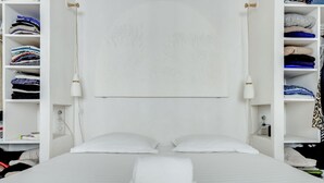 Ein weißes Bett mit zwei Kissen, einem weißen Kopfteil und einem weißen Kleiderschrank mit gefalteter Kleidung.