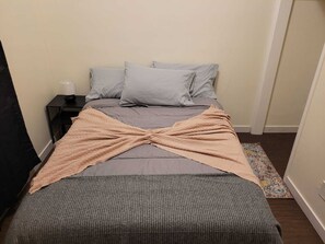 Un lit recouvert d’une couvre-lit grise, d’un plaid beige et de deux oreillers. Une petite table de chevet avec une lampe se trouve à côté du lit.
